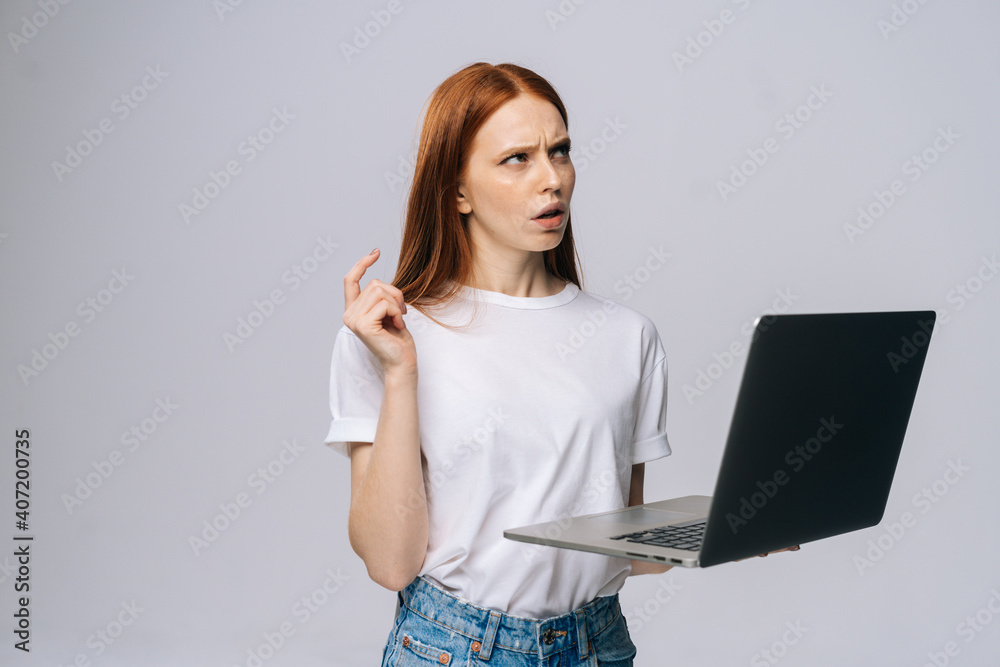 Thinking young woman student using laptop computer and looking away on isolated gray background. Pretty redhead lady model emotionally showing facial expressions in studio, copy space. Thinking young woman student using laptop computer and looking away on isolated gray background. Pretty redhead lady model emotionally showing facial expressions in studio, copy space.