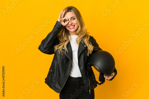 Young blonde caucasian biker woman holding helmet excited keeping ok gesture on eye.