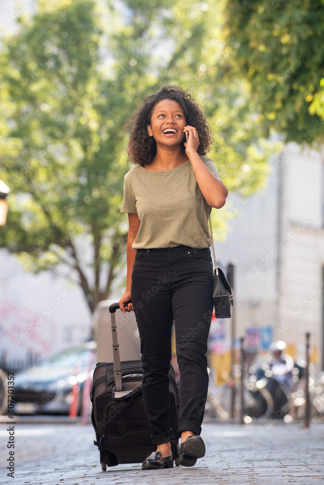 Full body happy young woman walking and talking with cellphone and suitcase in city Full body happy young woman walking and talking with cellphone and suitcase in city