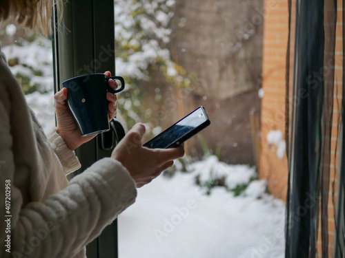 A shallow focus shot of a blonde woman with a smartphone and a mug watching snowing in front of the window