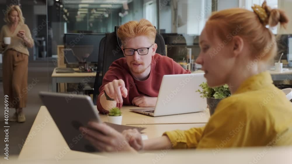 Medium close-up of two smiling red-haired office workers looking at tablet screen and discussing new project sitting at workplaces in bright and spacious office