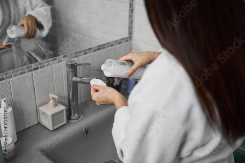 Woman in bathrobe using face cleanser indoors