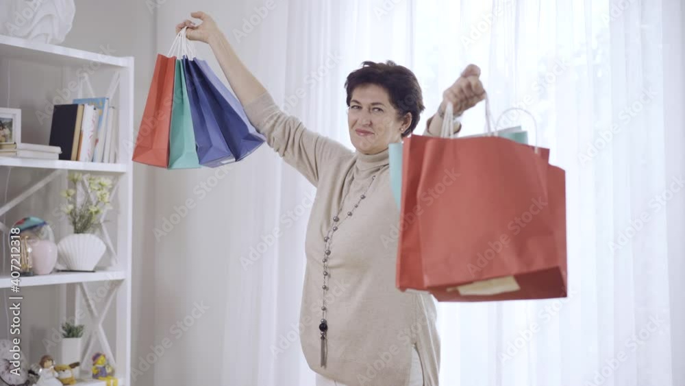 Middle shot portrait of happy middle-aged woman raising hands with shopping bags and smiling at camera. Positive confident Caucasian shopper posing indoors at home with purchases on Black Friday. Middle shot portrait of happy middle-aged woman raising hands with shopping bags and smiling at camera. Positive confident Caucasian shopper posing indoors at home with purchases on Black Friday.