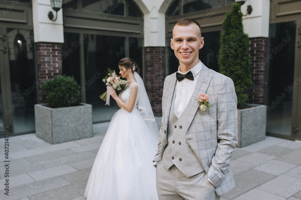 Naklejka premium Stylish groom in a gray suit and black bow-tie and the bride in a white dress with a bouquet in their hands are out of focus on the background of buildings in the city. Wedding portrait.