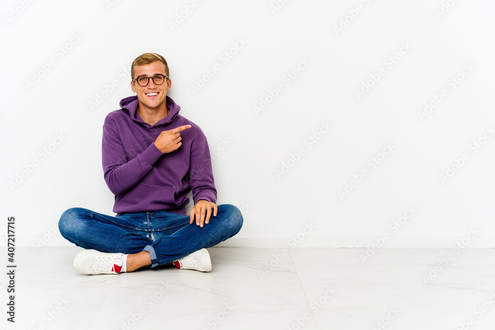Young caucasian man sitting on the floor smiling and pointing aside, showing something at blank space. Young caucasian man sitting on the floor smiling and pointing aside, showing something at blank space.