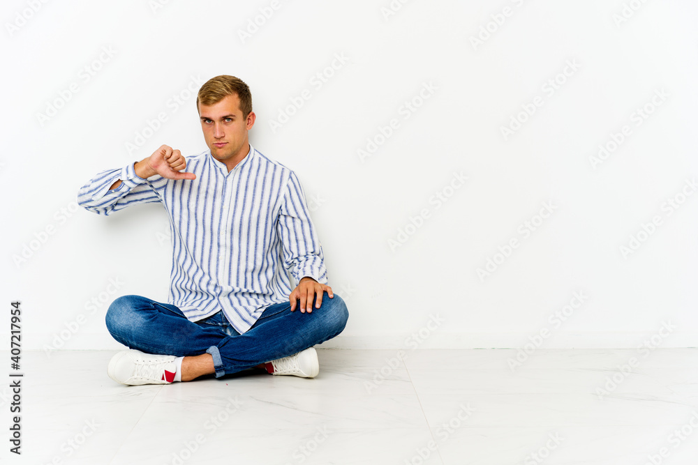 Young caucasian man sitting on the floor showing a dislike gesture, thumbs down. Disagreement concept. Young caucasian man sitting on the floor showing a dislike gesture, thumbs down. Disagreement concept.