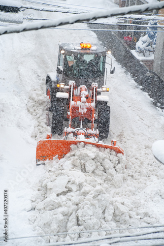Snowplow tractor removing snow in a narrow street during a heavy snowfall. Overhead shot with power lines in a foreground.