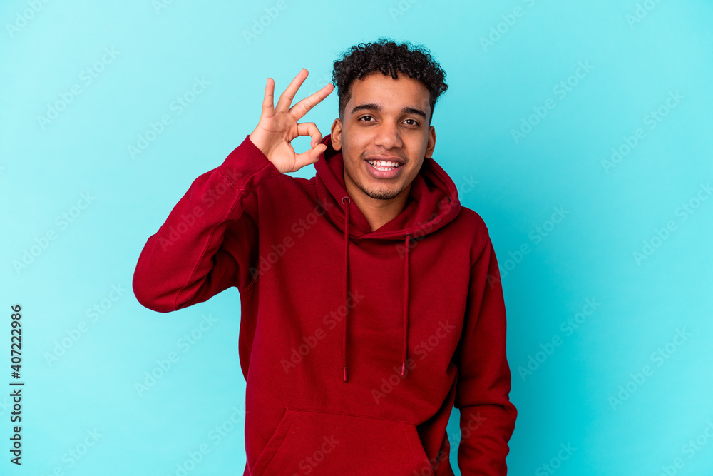 Young african american curly man isolated on blue winks an eye and holds an okay gesture with hand. Young african american curly man isolated on blue winks an eye and holds an okay gesture with hand.
