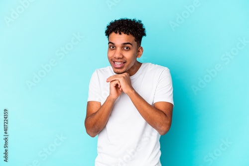 Young african american curly man isolated on blue keeps hands under chin, is looking happily aside.