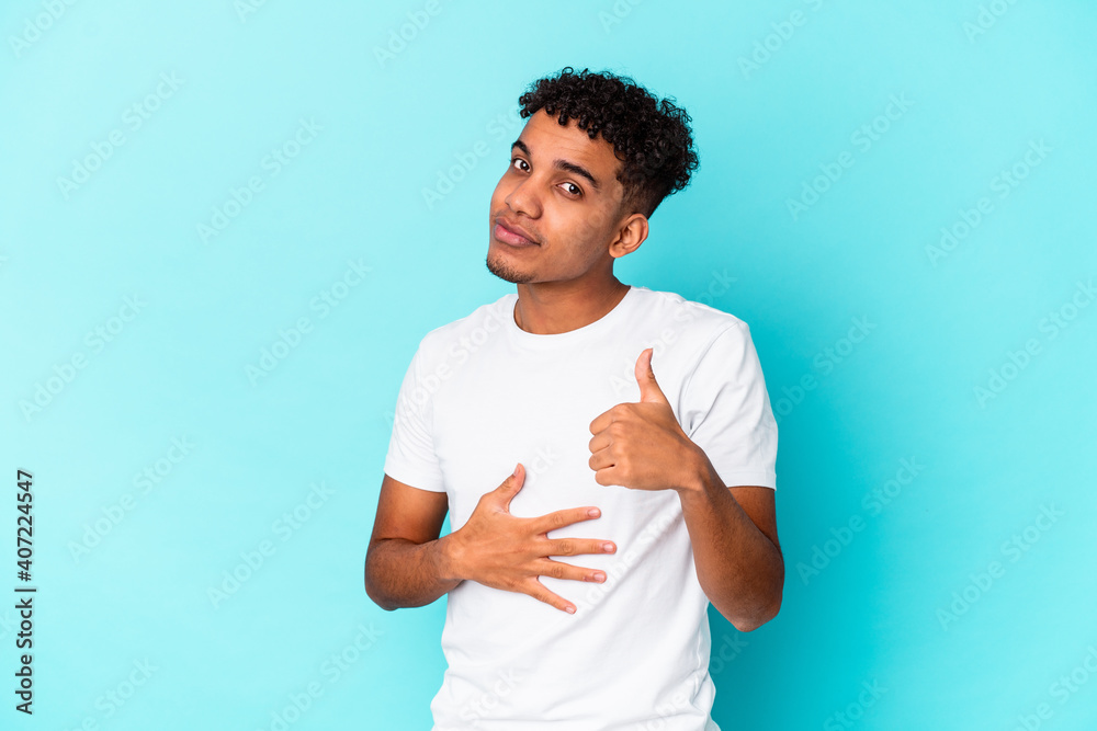 Young african american curly man isolated on blue touches tummy, smiles gently, eating and satisfaction concept. Young african american curly man isolated on blue touches tummy, smiles gently, eating and satisfaction concept.