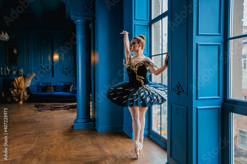 Ballerina poses in front of a large window, standing on the windowsill. Girl in a dark blue ballet tutu