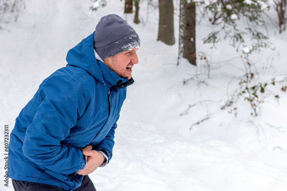 A man having a sudden stomach ache. Casual young adult man having stomach pain while walking in the snowy mountain. Man with stomach pain. Adult Caucasian man pressing on belly with painful expression