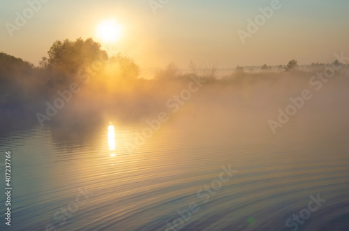 Sunrise over the foggy lake with the reflection of sun and trees in the water. Early fresh morning landscape. Mist on the water, forest silhouettes and the rays of the rising sun.