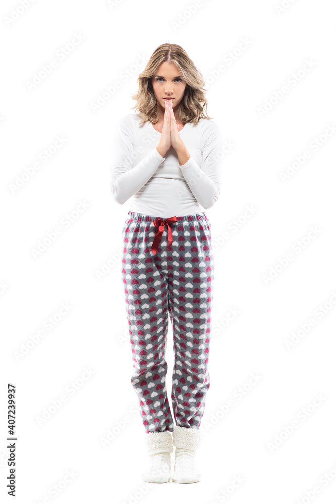 Young serious woman pleading staring at camera with praying hands in pajama. Full length portrait on white background.