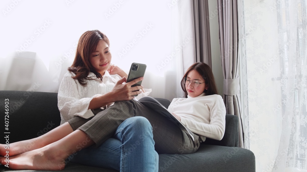 Lovely Asian couple lying on sofa play smartphone with a friend sitting and reading a book together daily holiday life of an Asian woman in the living room at home.