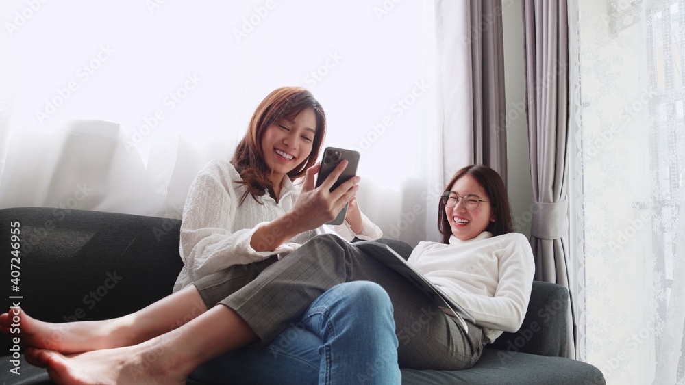 Lovely Asian couple lying on sofa play smartphone with a friend sitting and reading a book together daily holiday life of an Asian woman in the living room at home.