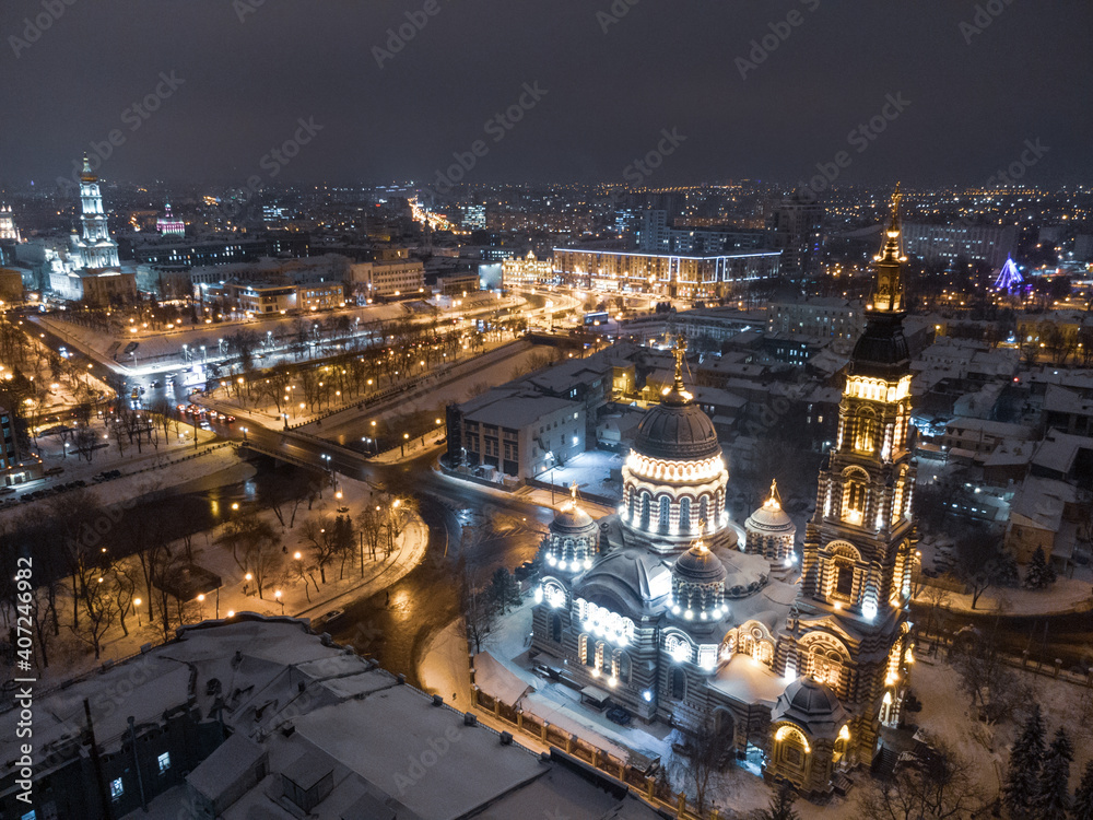 Fototapeta premium Holy Annunciation Cathedral illuminated in winter snowy night lights. Aerial view Kharkiv city orthodox church on riverbank, Ukraine. Side view from air.