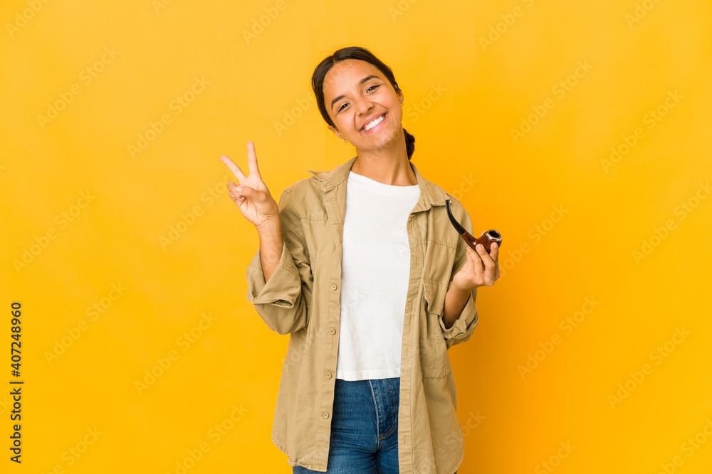 Young hispanic woman holding a smoking pipe joyful and carefree showing a peace symbol with fingers. Young hispanic woman holding a smoking pipe joyful and carefree showing a peace symbol with fingers.