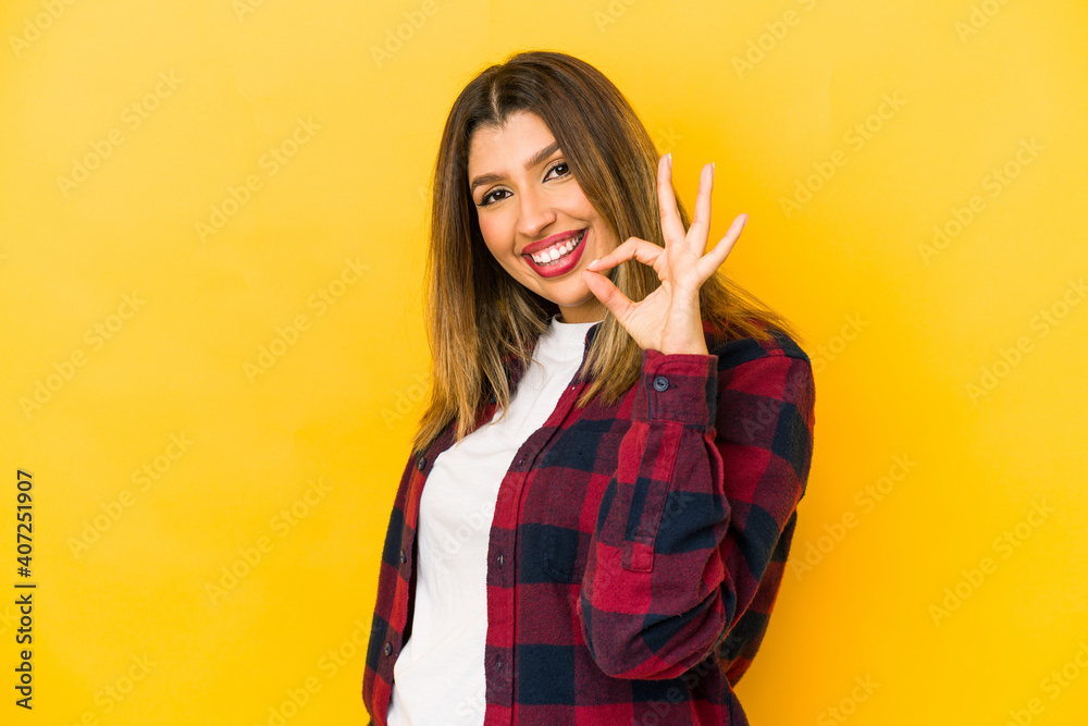 Young indian woman isolated on yellow background winks an eye and holds an okay gesture with hand. Young indian woman isolated on yellow background winks an eye and holds an okay gesture with hand.