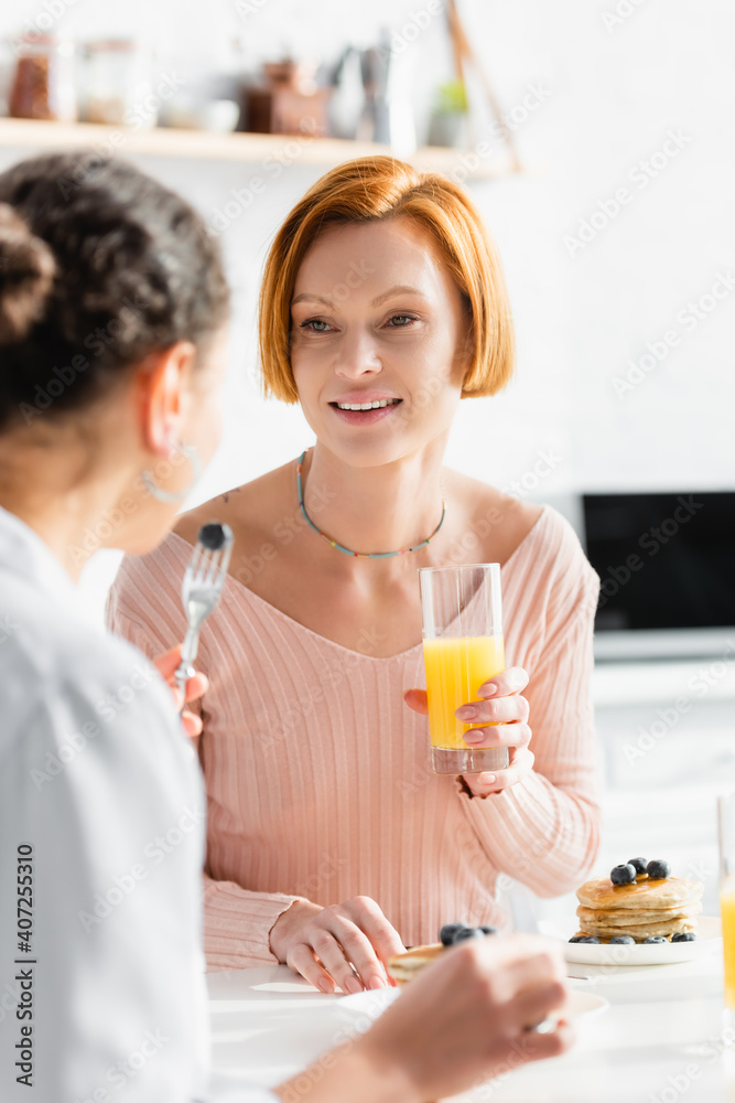 redhead lesbian woman holding orange juice near african american girlfriend with blueberry on fork, blurred foreground redhead lesbian woman holding orange juice near african american girlfriend with blueberry on fork, blurred foreground
