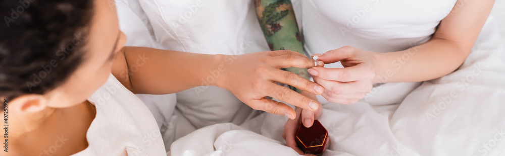 cropped view of woman putting wedding ring on finger of lesbian girlfriend in bedroom, blurred foreground, banner