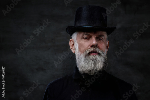 Dressed in old fashioned style clothing grandfather with top hat and gray hairs poses in dark background.