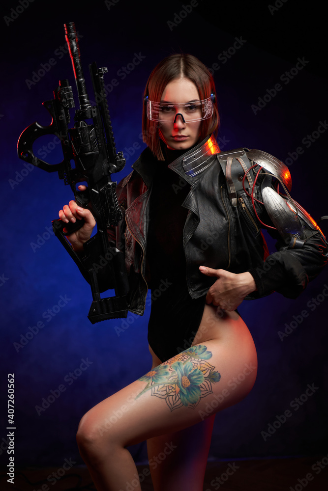 Attractive and sexy female soldier dressed in black jacket poses in dark background. Stylish woman with short haircut and glasses looks at camera holding futuristic rifle.