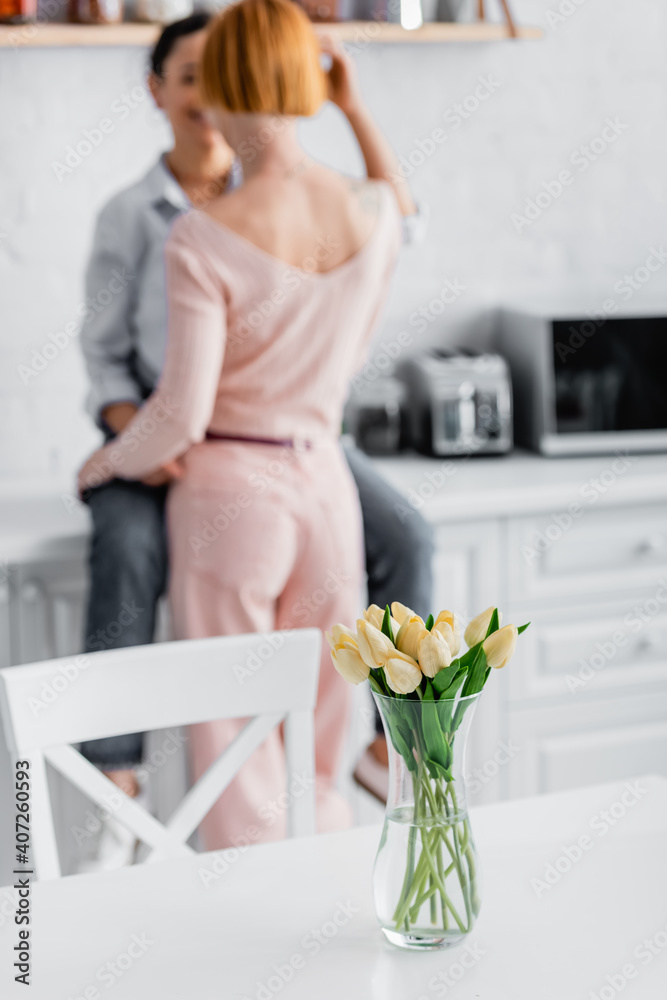 selective focus of tulips in vase on kitchen table near interracial lesbian couple on blurred background selective focus of tulips in vase on kitchen table near interracial lesbian couple on blurred background
