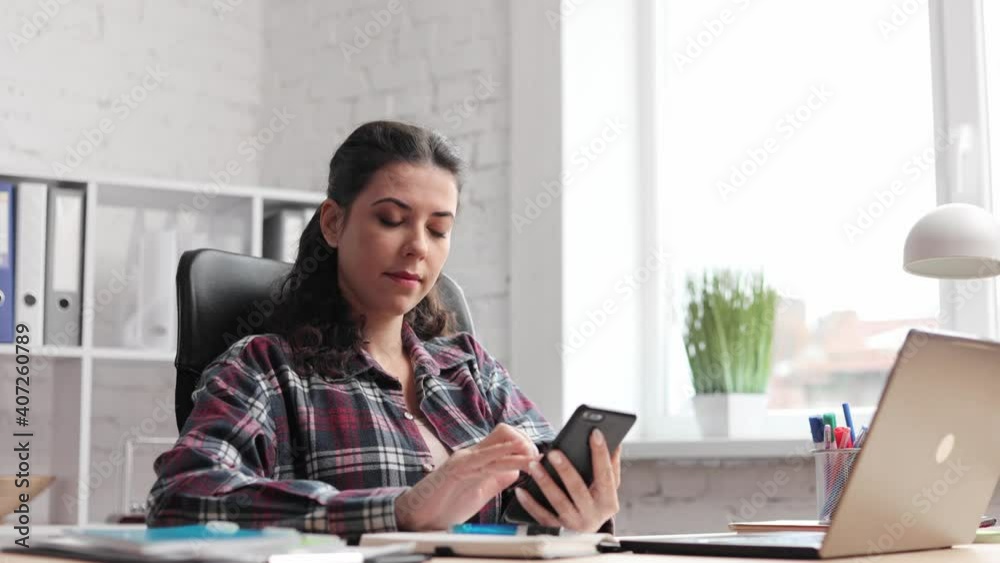Female watching and uses a cellphone in office. Freelance worker. Focused young woman tapping and scrolling on mobile phone. Close up. Portrait of curlyl brunette girl texting on smartphone, indoor. Female watching and uses a cellphone in office. Freelance worker. Focused young woman tapping and scrolling on mobile phone. Close up. Portrait of curlyl brunette girl texting on smartphone, indoor.