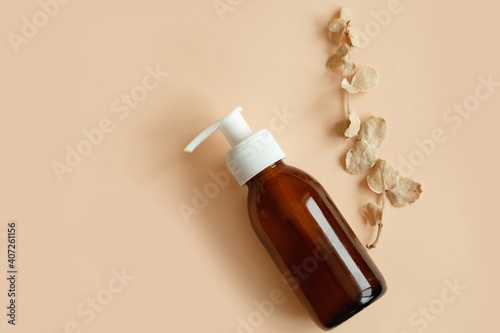 Brown glass bottle with a pump of cosmetic products framed by green leaf branches, beige background. Natural organic spa cosmetic beauty concept. Front view Mock up.