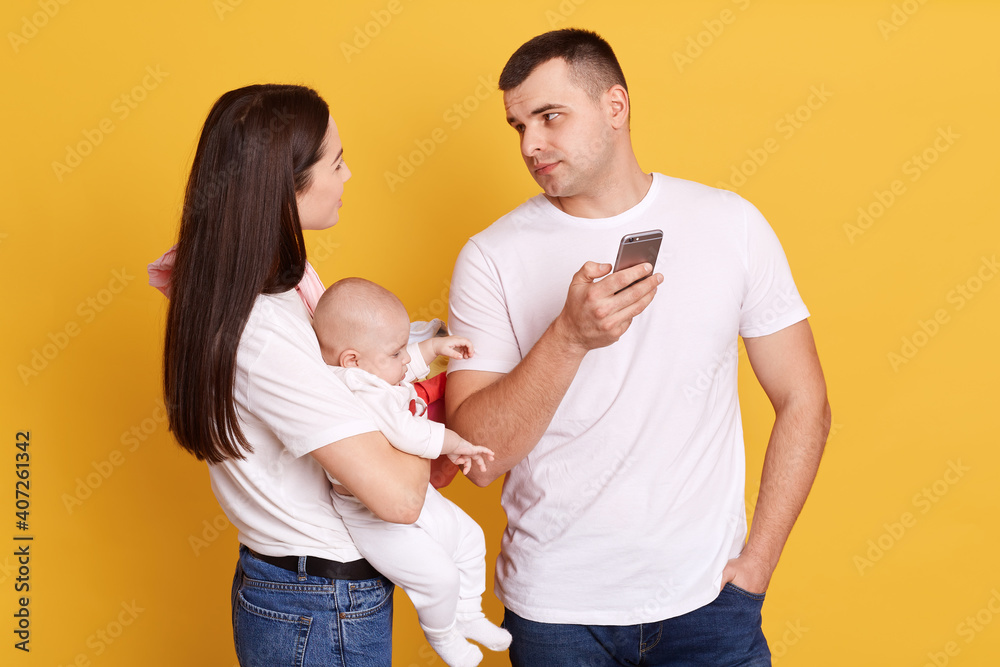 Young attractive mother with newborn daughter in hands looking at her husband and makes complaint because father does not help with house hold chores and child, posing isolated over yellow background. Young attractive mother with newborn daughter in hands looking at her husband and makes complaint because father does not help with house hold chores and child, posing isolated over yellow background.