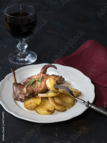 Roasted rabbit and potatoes on white plate and glass of red wine on dark table. Copy space.