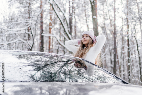 a young beautiful girl dressed in a winter light hat and a white sweater looks out of the car window in a winter snowy forest
