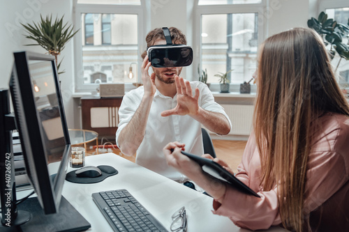 Caucasian guy weared with virtual reality glasses enjoys at work with his female colleague which uses tablet in office room. photo
