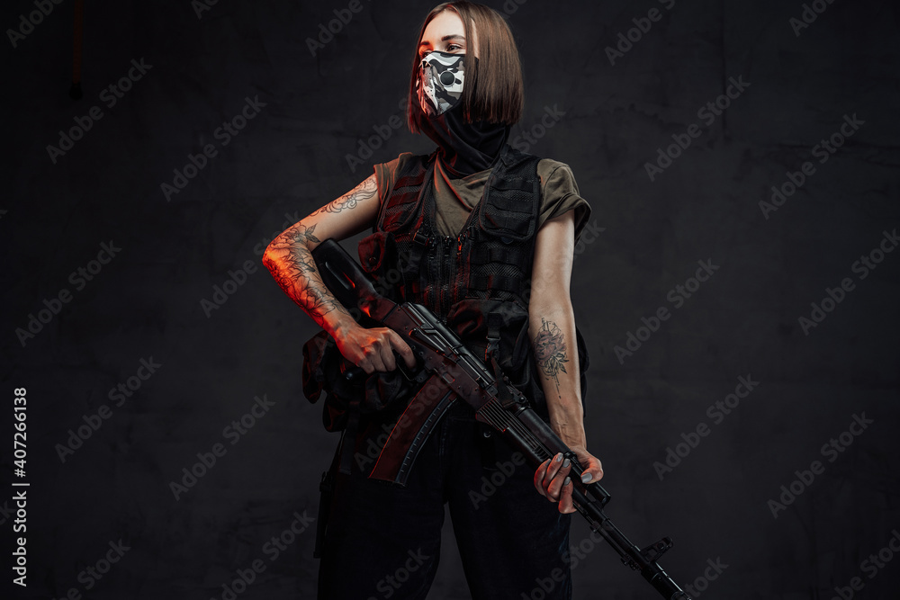 Dressed in dark armour and shirt female soldier with short haircut and weared with mask poses holding ak74 rifle.