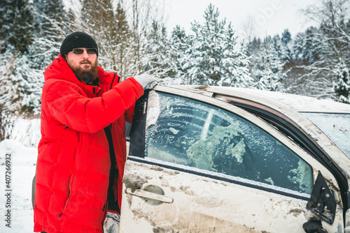 A man after an accident holds on to the door of a crashed car, on the side of the road in winter.