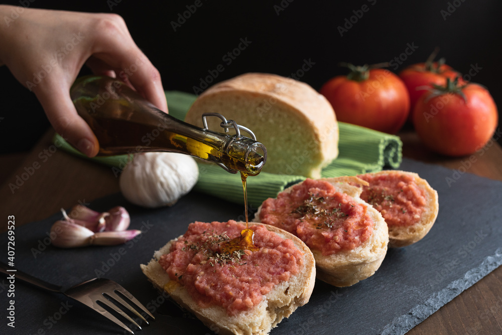 Mano echando aceite de oliva sobre unas tostadas de pan tumaca español con  pan, tomate, ajo, aceite de oliva y orégano con un fondo negro. Stock Photo  | Adobe Stock