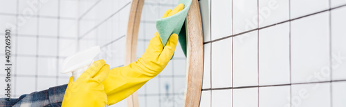 Cropped view of woman in rubber gloves cleaning mirror with rag and detergent in bathroom, banner