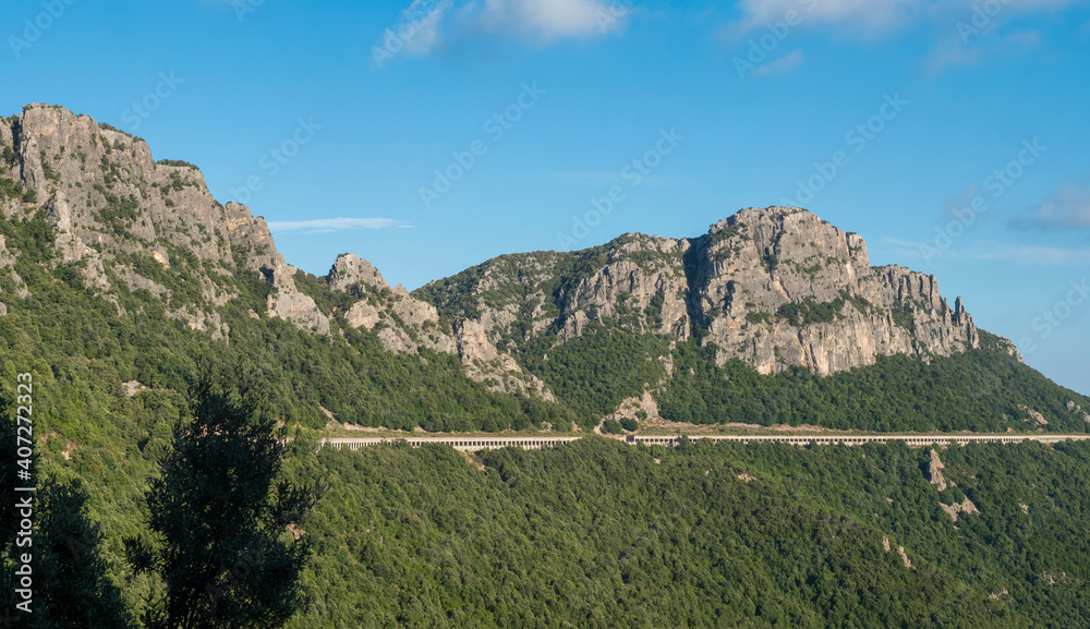 View of long open tunnel or viaduct with pillars and widows through limestone rock with green forest at Supramonte mountains. Genna Selole, Sardinia, Italy. Blue sky background