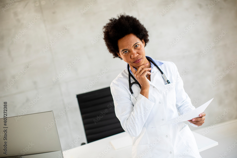 Female African American doctor wearing white coat with stethoscope standing by desk in office and looking at medical test results on paper Female African American doctor wearing white coat with stethoscope standing by desk in office and looking at medical test results on paper