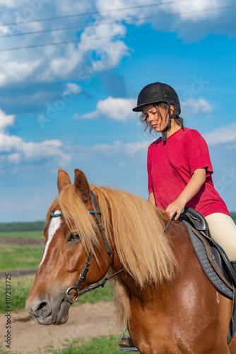 A young beautiful girl in a jockey cap and a red T-shirt rides a horse.
