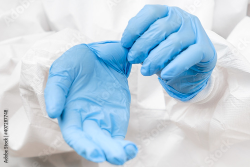 Doctor or nurse in protective PPE suit putting on rubber blue gloves before work with coronavirus patient or test samples or vaccination