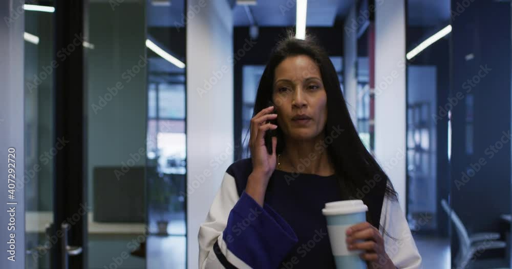 Mixed race businesswoman talking on smartphone holding coffee walking in office corridor Mixed race businesswoman talking on smartphone holding coffee walking in office corridor