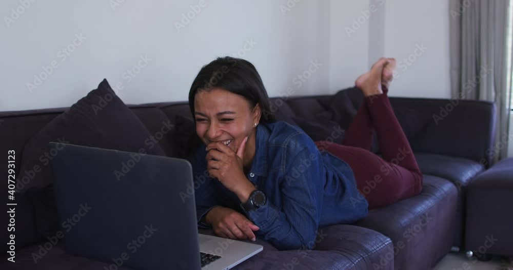 Mixed race woman lying on couch using laptop and laughing Mixed race woman lying on couch using laptop and laughing