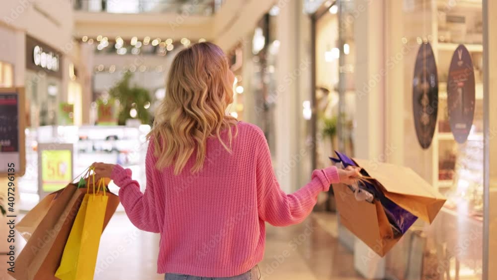 Portrait of happy attractive young woman in headphones walks around shopping mall. Smiling girl in pink sweater with gift colored bags looking to the camera. Pretty happy shoper.