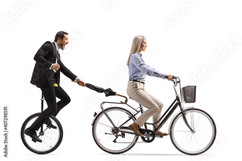 Man in a suit and tie holding an umbrella and riding a tricycle behind a woman on a bicycle