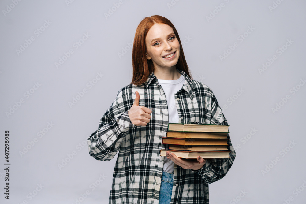 Beautiful young woman college student holding book and showing thumb up gesture looking at camera on gray isolated background. Pretty redhead lady model emotionally showing facial expressions. Beautiful young woman college student holding book and showing thumb up gesture looking at camera on gray isolated background. Pretty redhead lady model emotionally showing facial expressions.