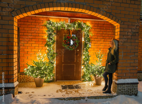 a woman leaning against the wall at the entrance to a house decorated with Christmas decor. ate in flowerpots, decorated with a garland and a Christmas wreath on the door photo