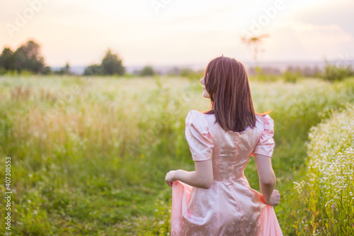 Beautiful happy woman with dark hair dancing in the wildflowers field holding the bottom of the long pink dress on a warm summer evening. Joyfull summer walking. 