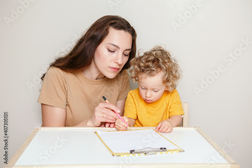 Mom drawing on paper together with baby boy toddler. Mother teaching kid to hold marker, pencil correctly. Early age kids education development learning. Family candid life at home.